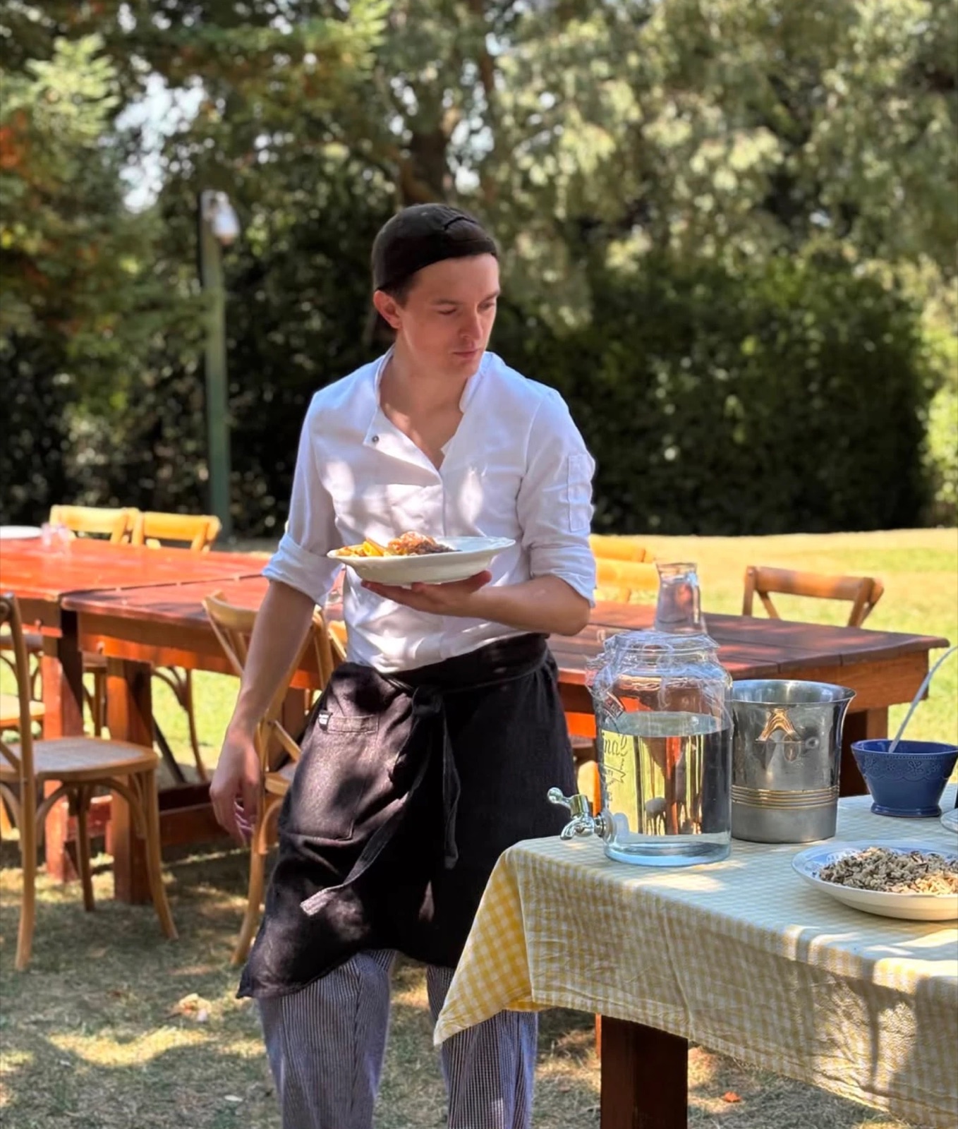 Arthur en veste de cuisine, assiette à la main, au service d'un repas dans un jardin